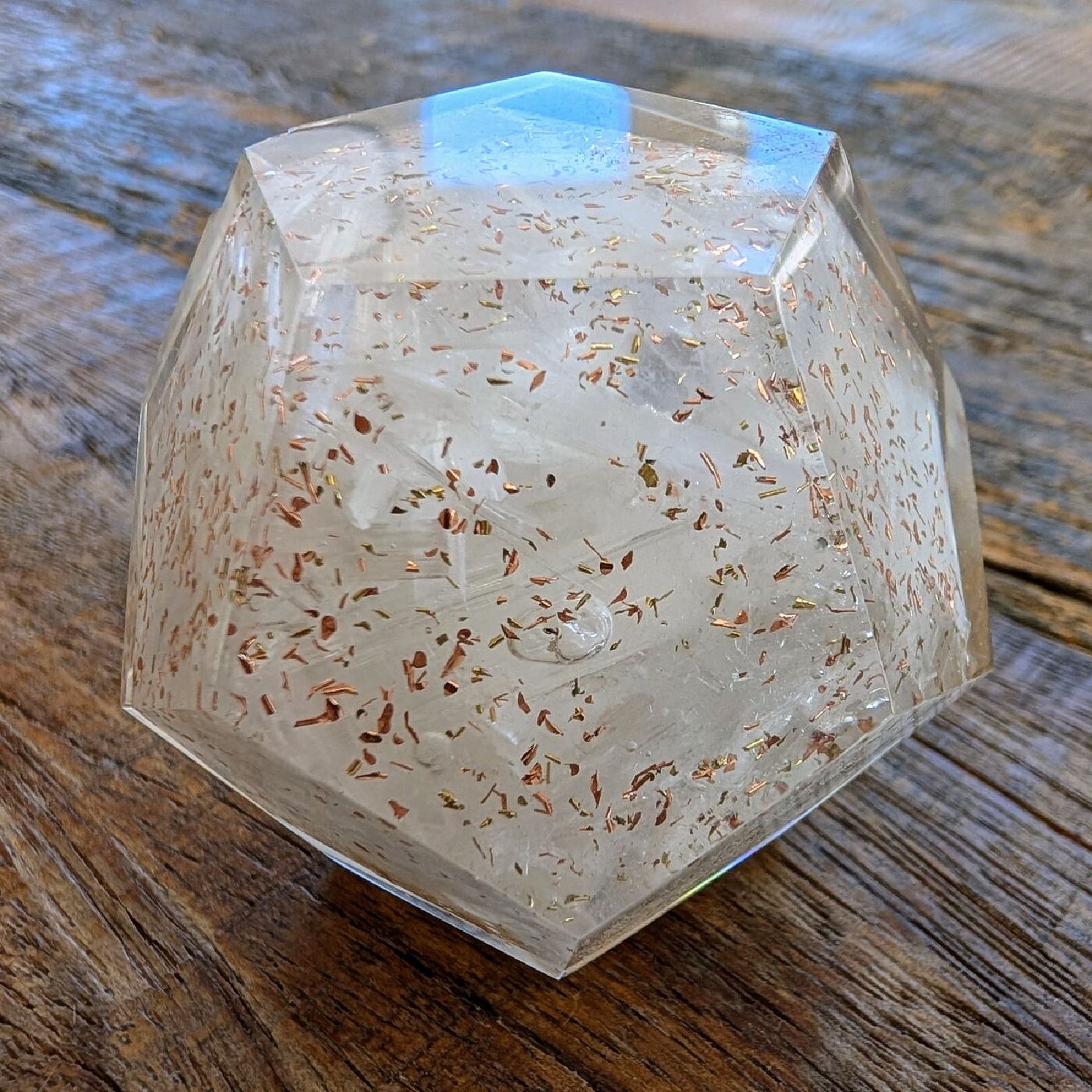A clear twelve-sided crystal with copper flecks inside, on a wooden rail in evening sun, a garden behind it